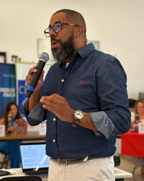 Marcelo Sales falando ao microfone em evento, com notebook aberto ao lado